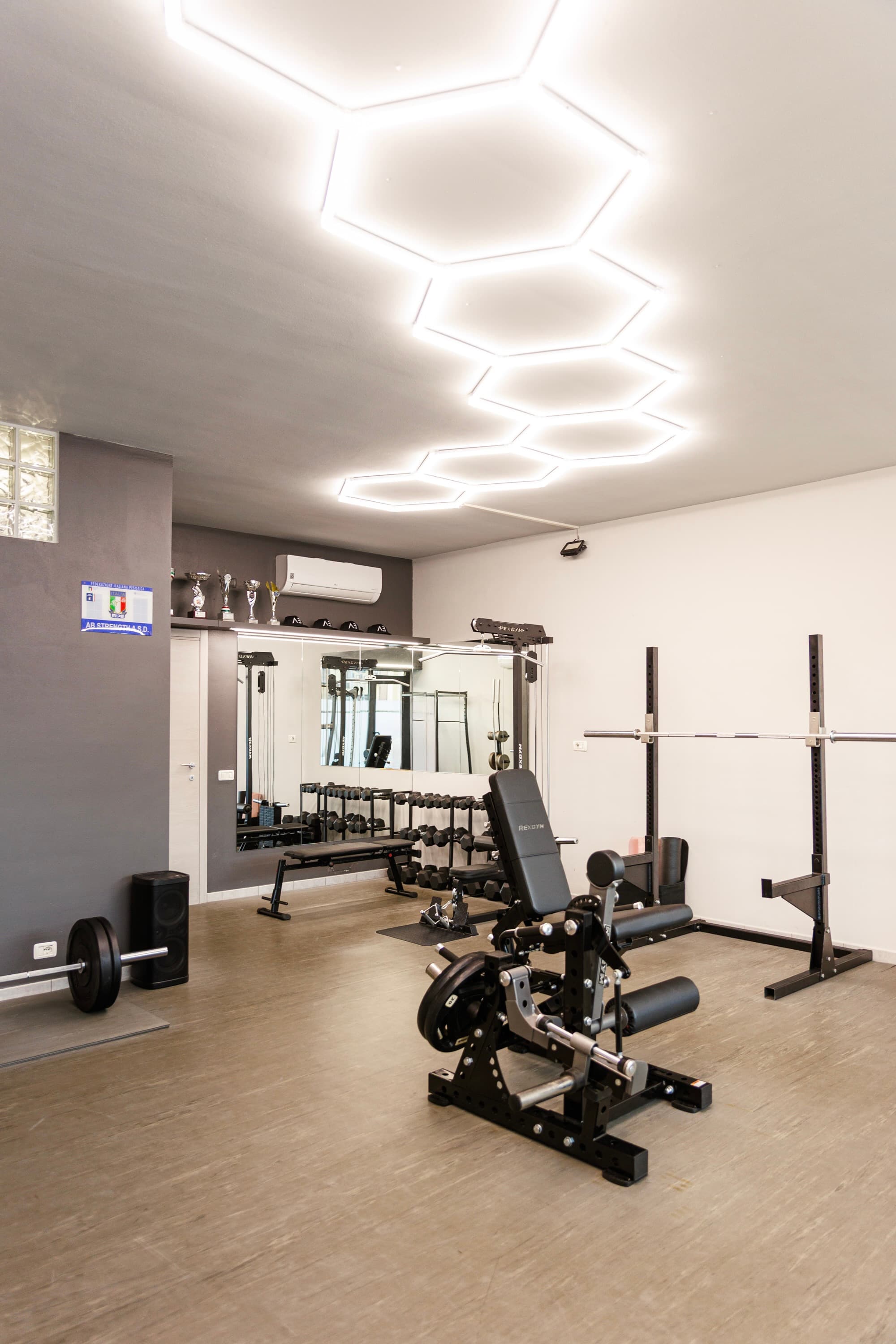 La sala pesi sotto le luci a esagono, con lo specchio, i rack e le macchine